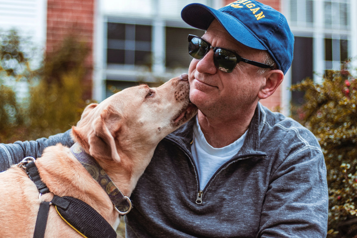 A man wearing black sunglasses and a blue baseball hat wraps an arm around a yellow labrador retriever as he gets a kiss from the dog.